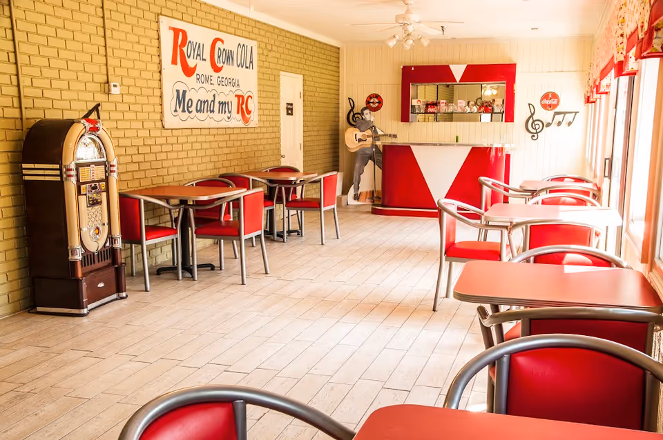 A retro-style dining area with red chairs and tables, a vintage jukebox on the left, a red and white counter at the back, and wall decorations including a large Royal Crown Cola sign and musical notes. The room has light-colored wooden flooring and large windows on the right side letting in natural light.