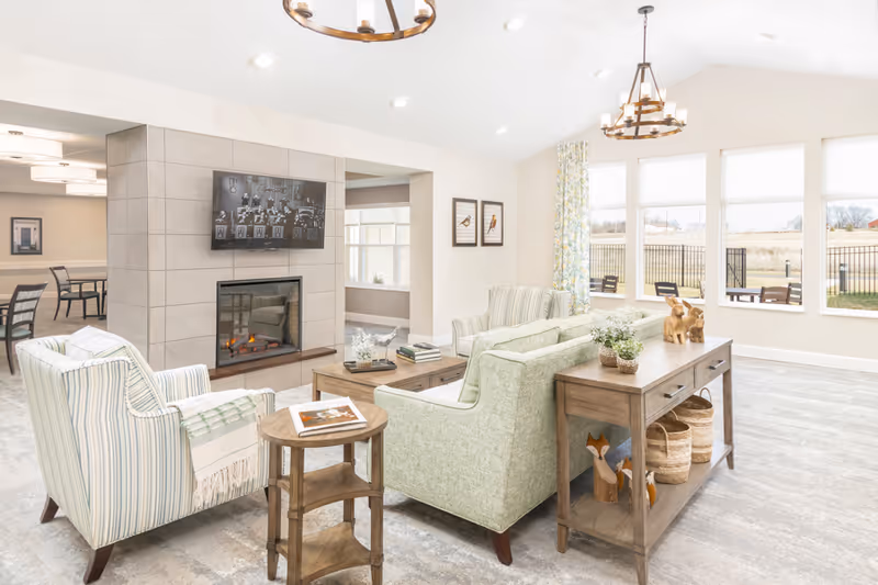 A bright and spacious living room area with large windows letting in natural light. The room features a fireplace with a mounted flat-screen TV above it, two armchairs, a sofa, a wooden coffee table, and a console table with decorative items and baskets underneath. Outside the windows, there is a fenced outdoor area with patio furniture.