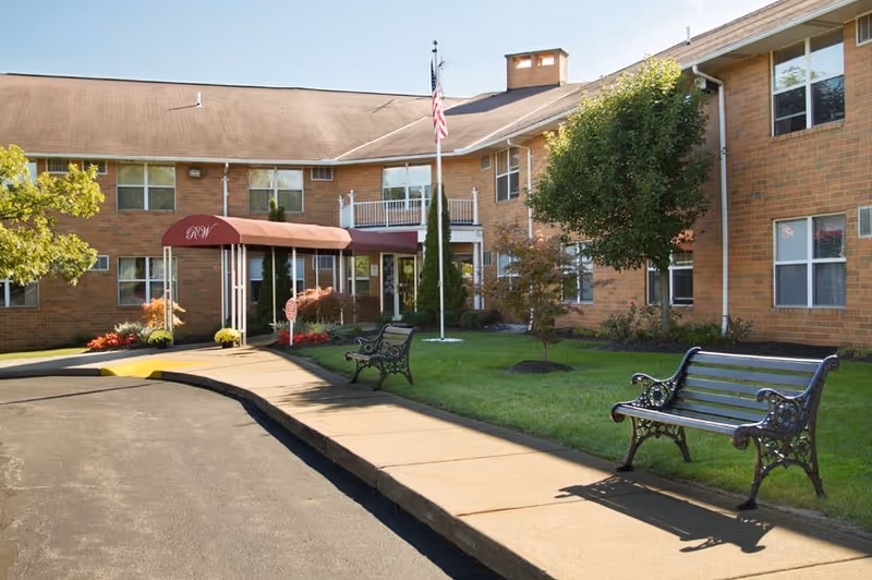 Exterior view of a two-story brick building with a maroon awning entrance labeled 'RW'. There are two black metal benches on a sidewalk leading to the entrance, a flagpole with an American flag, and some small trees and shrubs on a well-maintained lawn.