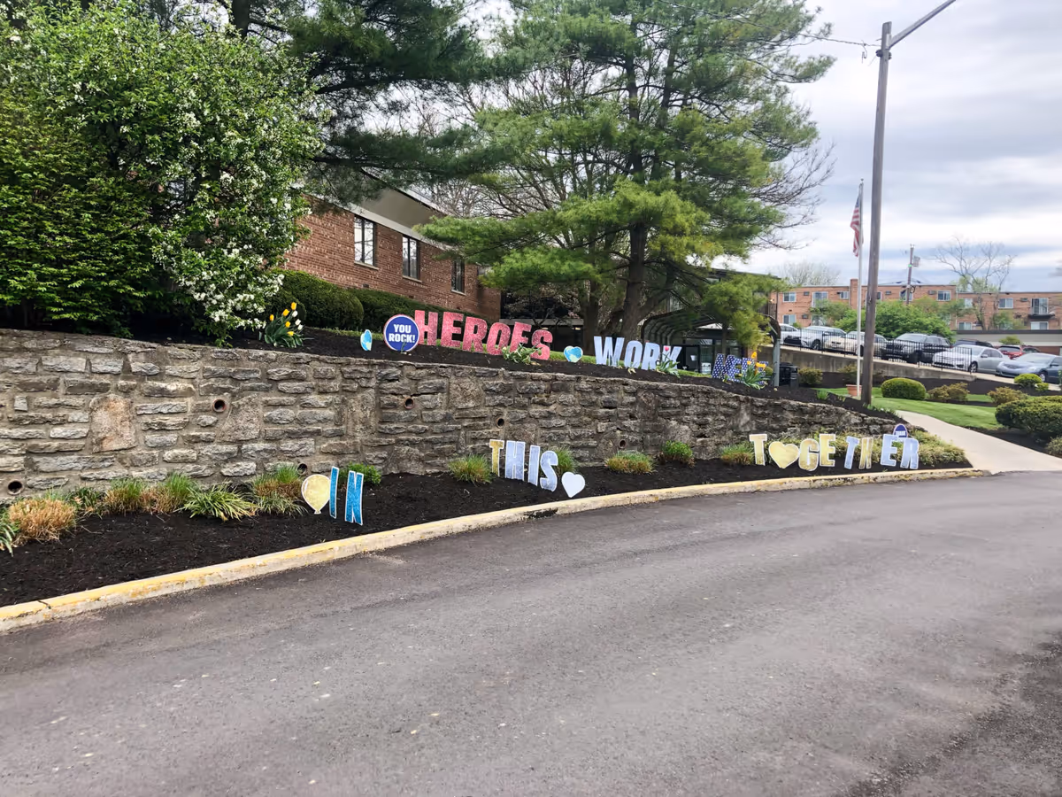 Driveway and stone retaining wall in front of a brick building with trees and colorful yard signs reading messages like "HEROES", "WORK" and "TOGETHER".