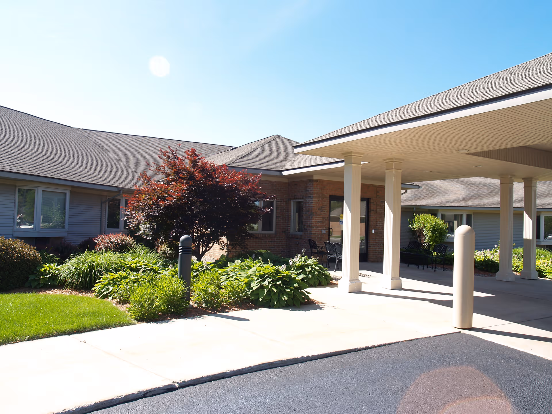 Exterior view of Seminole Shores Living Center showing a covered entrance with white pillars, a brick and siding building, landscaped greenery including bushes and a tree with red leaves, and a clear blue sky.
