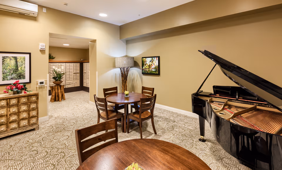 Communal lounge with round wooden tables and chairs, a grand piano, decorative lamp, and mailboxes in the background.