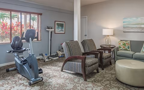 A room with exercise equipment including a stationary bike and a scale near a window with blinds. The room also has two patterned armchairs with a small wooden table between them, a green sofa with floral cushions, a round ottoman, a table lamp, and a landscape painting on the wall.