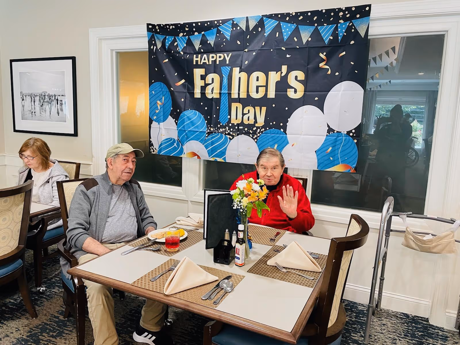 Two elderly men sitting at a dining table in a senior living facility with a 'Happy Father's Day' banner hanging on the wall behind them. One man is wearing a gray shirt and cap, the other is in a red sweater waving at the camera. The table is set with placemats, napkins, utensils, and a small flower arrangement. Another elderly woman is seated at a nearby table.