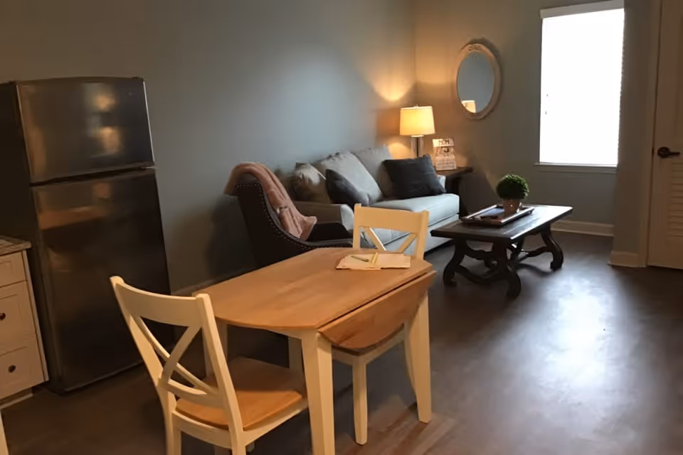 Small open living area with a wooden dining table and chairs in the foreground, a sofa and coffee table by a window, and a stainless refrigerator on the left.