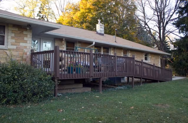Exterior view of a single-story senior living facility building with a long wooden ramp and railing leading to the entrance. The building has a stone facade, a chimney, and is surrounded by green grass and trees with autumn foliage.