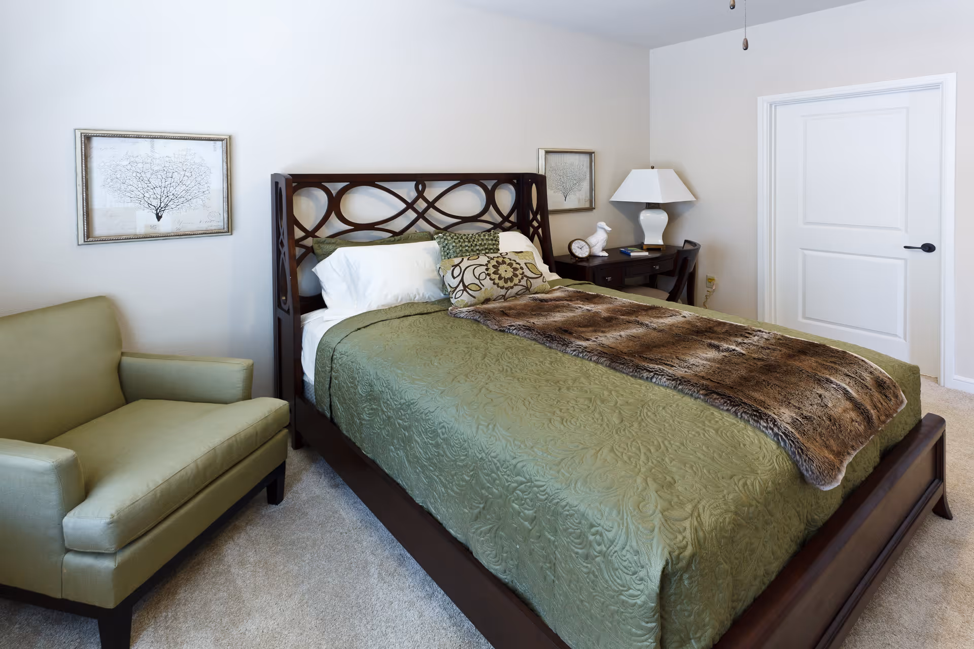 A bedroom with a dark wooden bed frame, green bedspread, decorative pillows, and a brown fur throw blanket. There is a green armchair to the left, a nightstand with a white lamp, clock, and decorative items to the right, and two framed pictures on the wall. A closed white door is visible in the background.