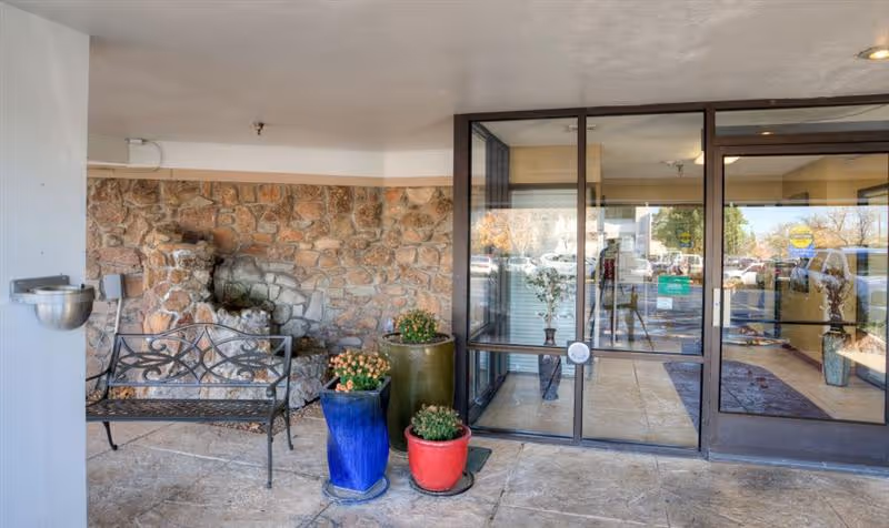 Covered entrance area of a facility with a stone wall, a black metal bench, a water fountain on the wall, and three potted plants in blue, green, and red pots. To the right, there is a glass door entrance with reflections of parked cars and trees outside.