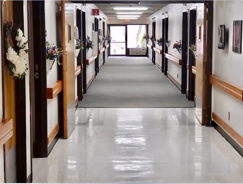 A clean, well-lit hallway in a senior living facility with white walls, wooden door frames, and handrails on both sides. Several doors line the corridor, each decorated with floral arrangements. The floor transitions from shiny white tiles to carpet as the hallway extends toward a glass exit door at the far end.