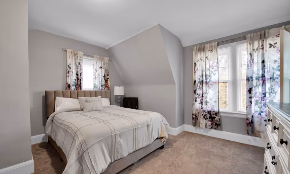 A cozy bedroom with a large bed featuring a beige headboard and light-colored bedding. The room has light gray walls, carpeted floor, and two windows with floral patterned curtains allowing natural light to enter. A white dresser with black knobs is visible on the right side, and a small lamp is placed near the bed.