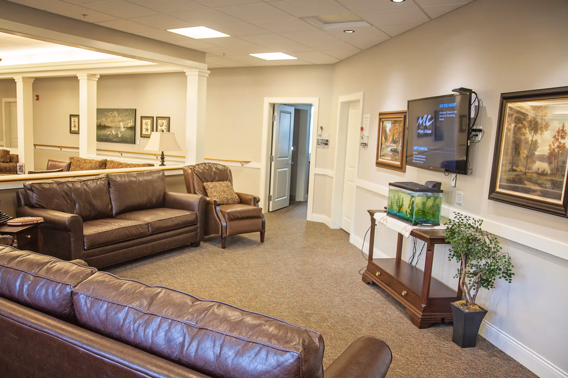 A bright lounge area with brown leather sofas and an armchair, a wall-mounted TV, an aquarium on a wooden table, and framed artwork on the walls.