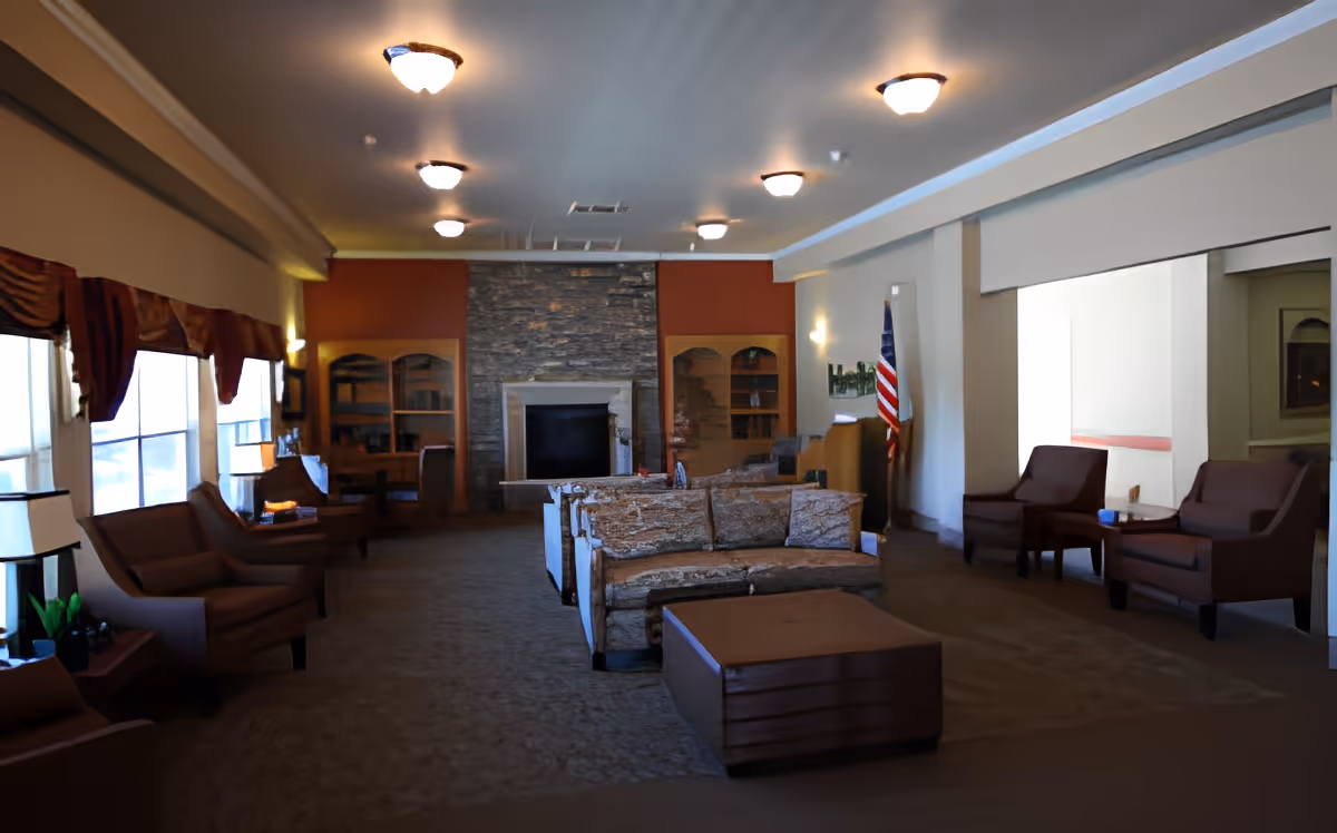 A spacious living room area with multiple armchairs and sofas arranged around a central coffee table. The room features a stone fireplace on the far wall flanked by wooden cabinets with glass doors. Large windows on the left side allow natural light to enter, and an American flag is positioned near the right wall. The ceiling has several light fixtures providing illumination.