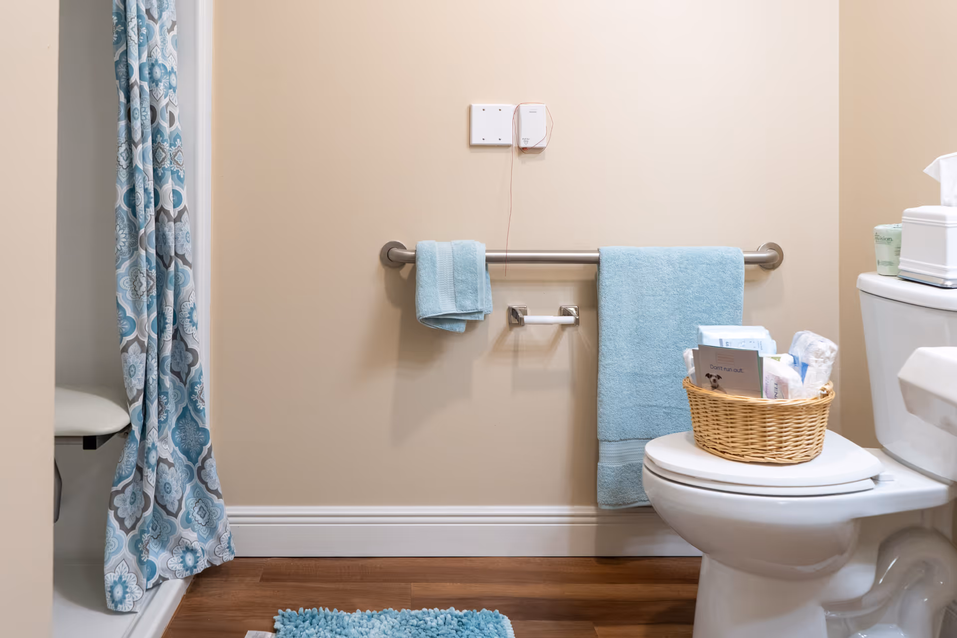A bathroom with beige walls and wood flooring featuring a white toilet with a wicker basket on the closed lid containing various items. There is a blue bath mat on the floor and a blue and white patterned shower curtain partially visible on the left. A silver grab bar is mounted on the wall with two blue towels hanging on it, and an empty toilet paper holder is attached below the grab bar.