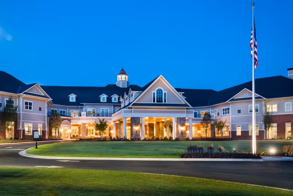 Exterior view of Sterling Estates of West Cobb Retirement Community building at dusk, featuring a large entrance with columns, well-lit windows, manicured lawn, and an American flag on a flagpole in front.