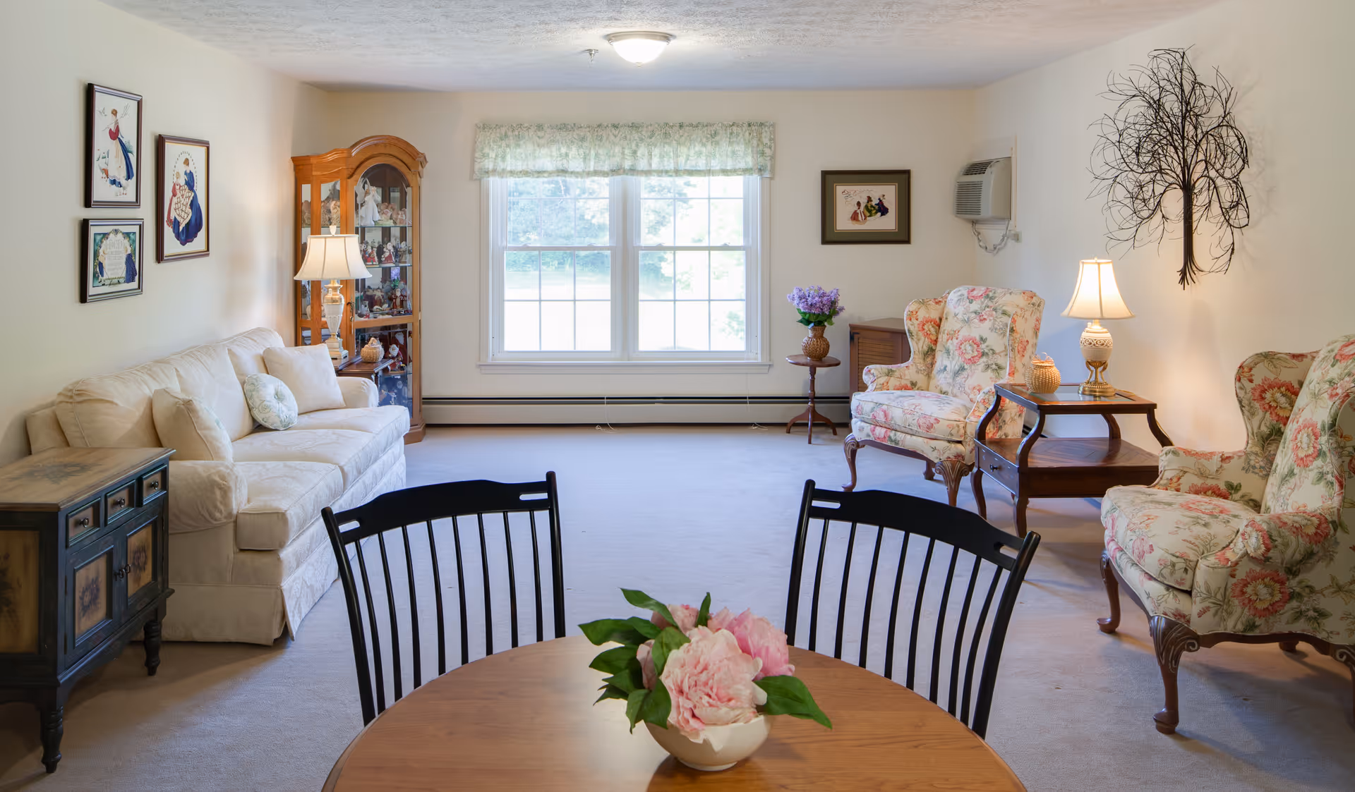 A cozy living room in a senior living community featuring a white sofa with cushions on the left, two floral upholstered armchairs on the right, a wooden coffee table with a lamp between the armchairs, a round wooden dining table with black chairs in the foreground, and a large window with floral valance in the center background. The room is decorated with framed pictures on the walls, a wooden display cabinet, a small side table with a flower vase, and a metal tree wall art.