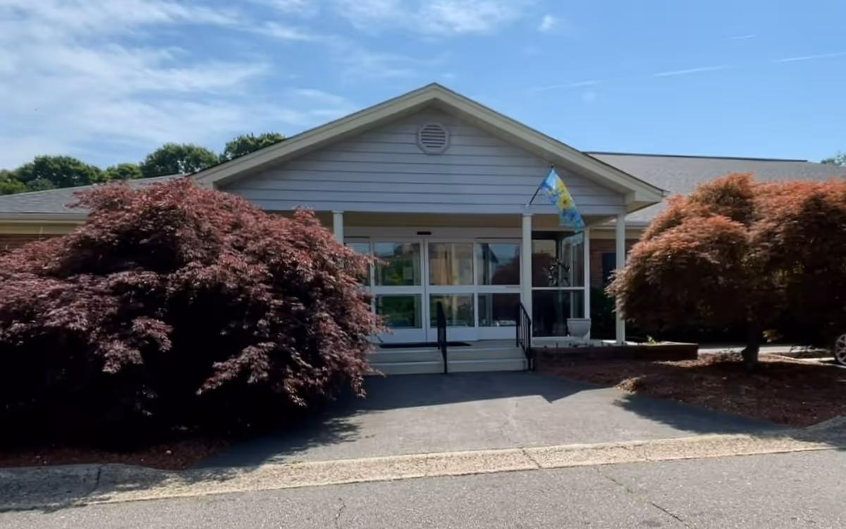 Front exterior view of a single-story building with a peaked roof, large windows, and a small porch with steps and railings. There are two large bushes with reddish leaves on either side of the entrance, and a flag with a sunflower design is hanging near the door. The sky is partly cloudy.