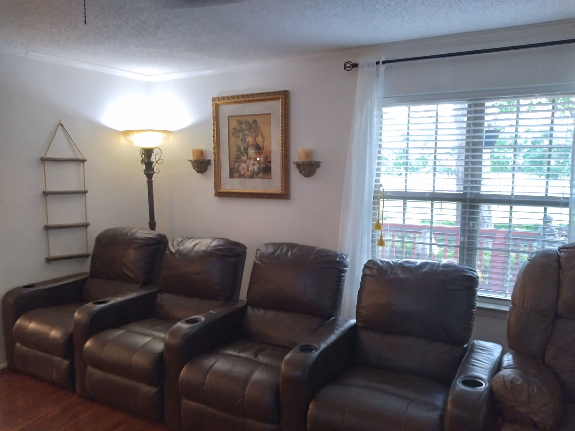 A cozy living room area with four dark brown leather recliner chairs arranged side by side. There is a tall floor lamp with a glowing light in the corner, a framed floral painting on the wall, two wall-mounted candle holders with candles, and a window with white blinds and sheer curtains letting in natural light.