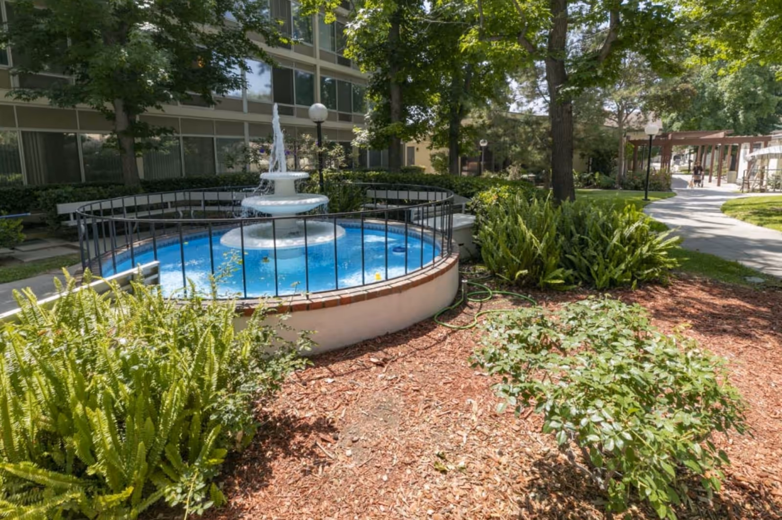 Outdoor garden area at Mission Care & Rehabilitation Center featuring a circular water fountain surrounded by a black metal fence, lush green plants, trees, and a paved walkway with lamp posts and a pergola in the background.