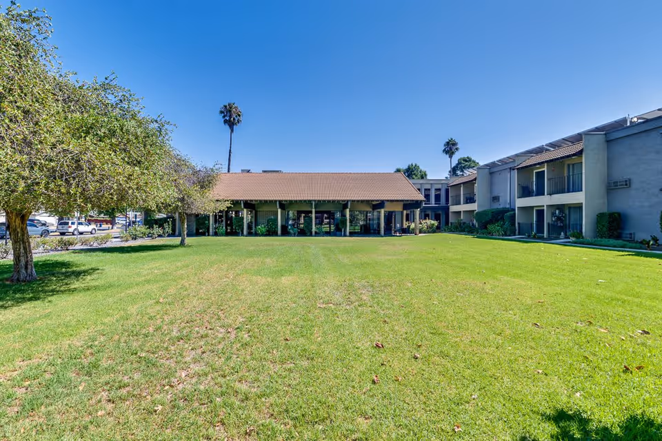A large green lawn with a few scattered leaves in front of a senior living facility building under a clear blue sky. The building has a tiled roof and multiple windows, with some trees and shrubs around the lawn. Two tall palm trees are visible in the background.