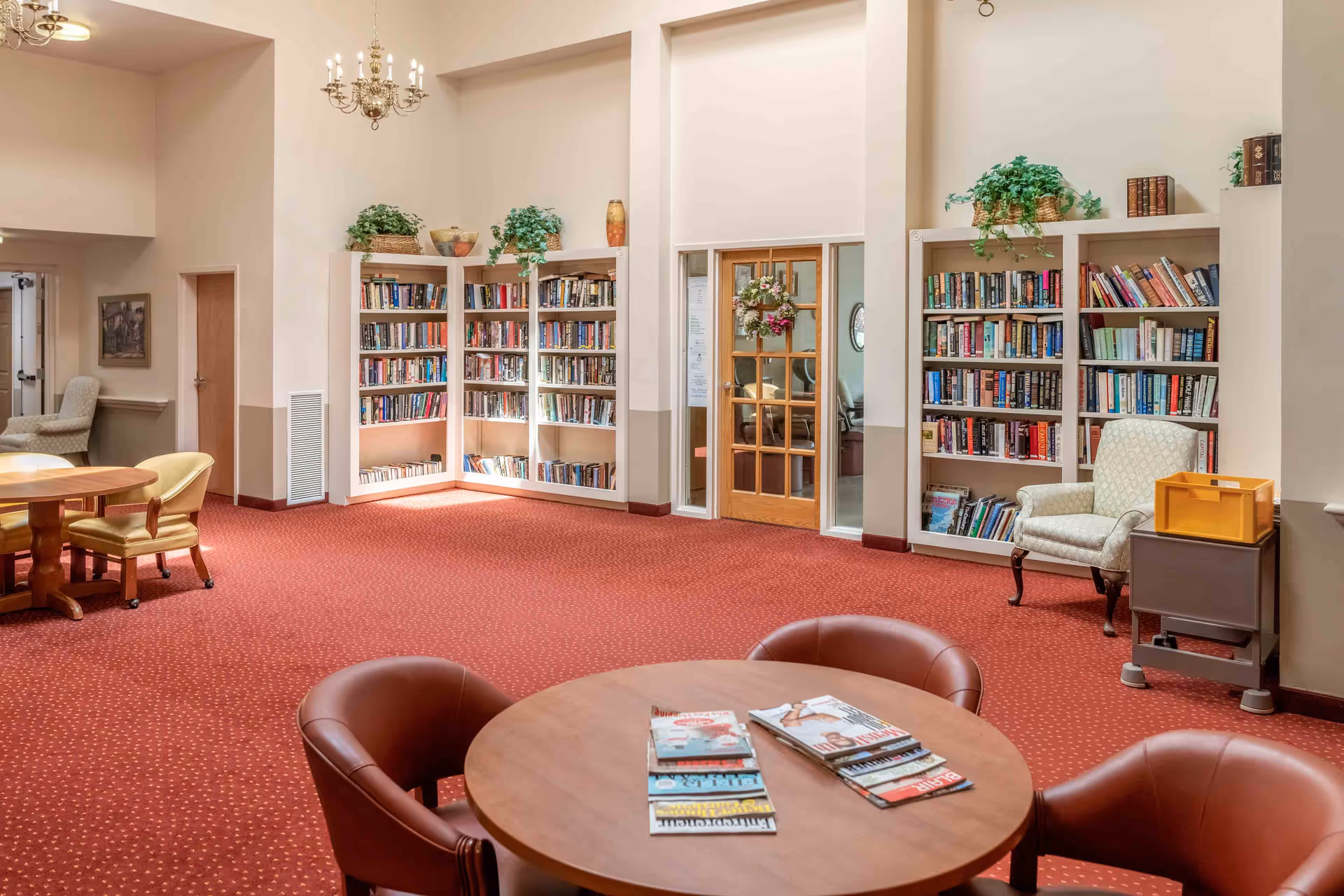 Bright communal library/lounge with bookshelves, round tables topped with magazines, upholstered chairs and red carpeting.