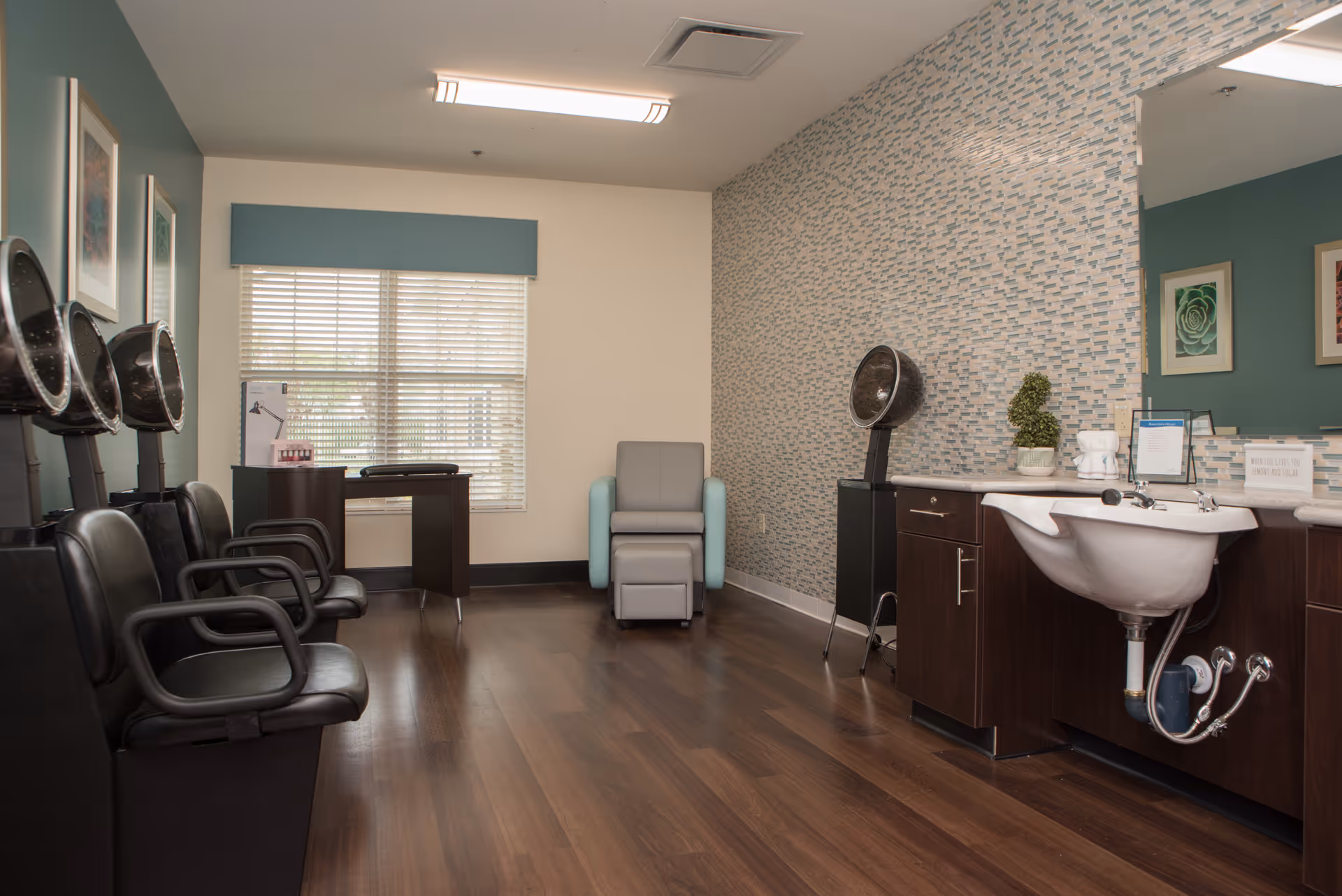 Bright salon room with multiple hair-drying chairs on the left, a reclining chair in the center, and a shampoo sink and counter on the right against a patterned wall.
