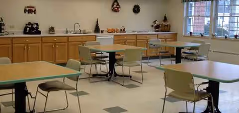 Bright dining room with pale green tables and chairs, along a counter and wall cabinets.