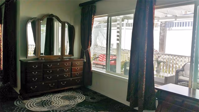 A room with a large wooden dresser featuring multiple drawers and a three-panel mirror. The room has dark patterned carpet and dark curtains framing a large window that looks out onto a patio with outdoor furniture and a white fence.