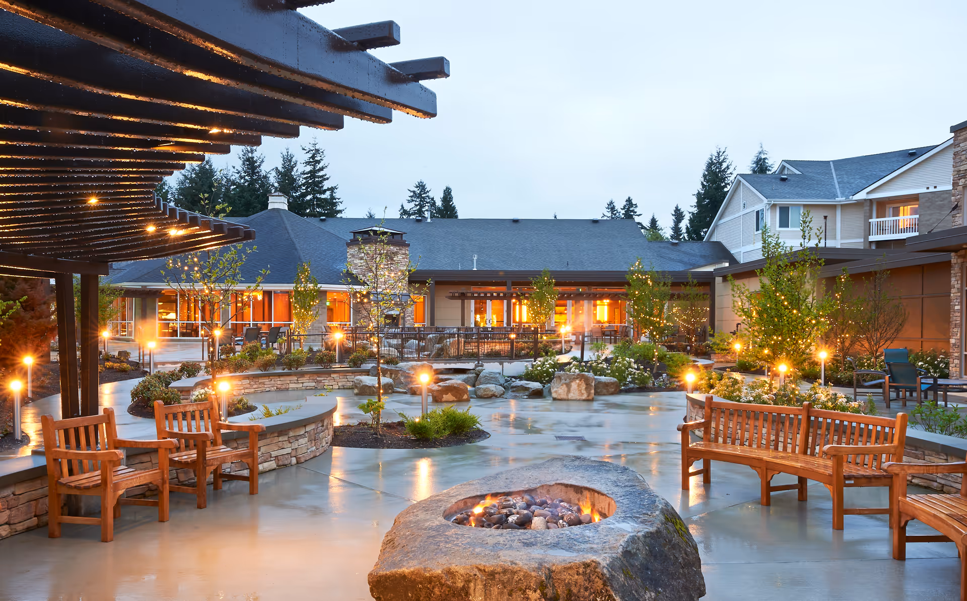 Well-lit outdoor courtyard with wooden benches, a stone fire pit, a pergola, and pathways in front of a residential building.