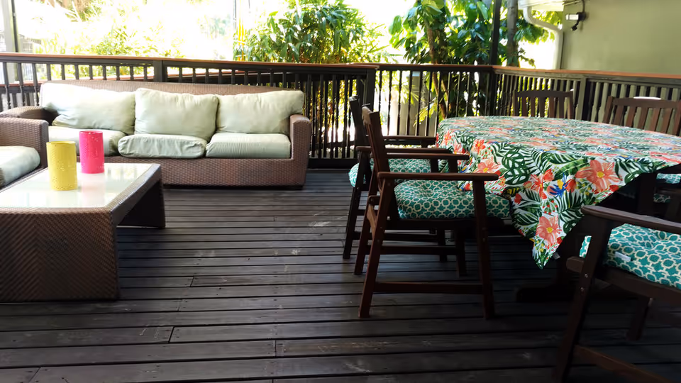 Outdoor patio area with a wicker sofa with light green cushions, a glass-top wicker coffee table with two colorful candle holders, and a wooden dining table covered with a floral tablecloth surrounded by wooden chairs with blue patterned cushions. The area is enclosed by a railing and surrounded by greenery.
