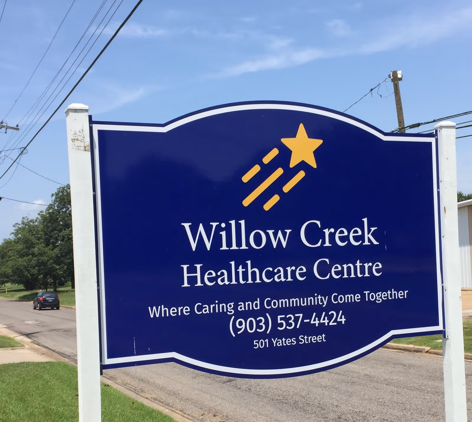 Outdoor sign for Willow Creek Healthcare Centre with a blue background and yellow shooting star graphic, displaying the tagline 'Where Caring and Community Come Together', phone number (903) 537-4424, and address 501 Yates Street.