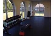 Small fitness room with a dumbbell rack, aerobic step bench, TV, and arched windows overlooking a lawn and neighboring building.