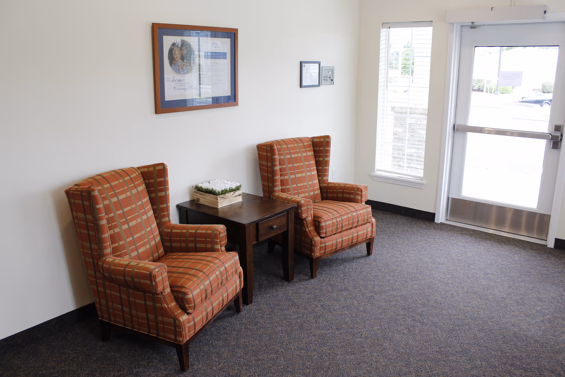 A small seating area with two orange plaid armchairs separated by a dark wooden side table with a decorative box on it. The area has a carpeted floor, white walls, a framed picture on the wall, a window with blinds, and a glass door leading outside.