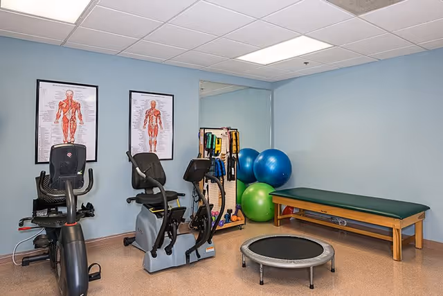 A small exercise room with two stationary recumbent bikes, a mini trampoline, a wooden exercise bench with a green cushion, several large exercise balls, and a rack holding various resistance bands and weights. The walls are light blue with two anatomical muscle charts hanging on one wall.