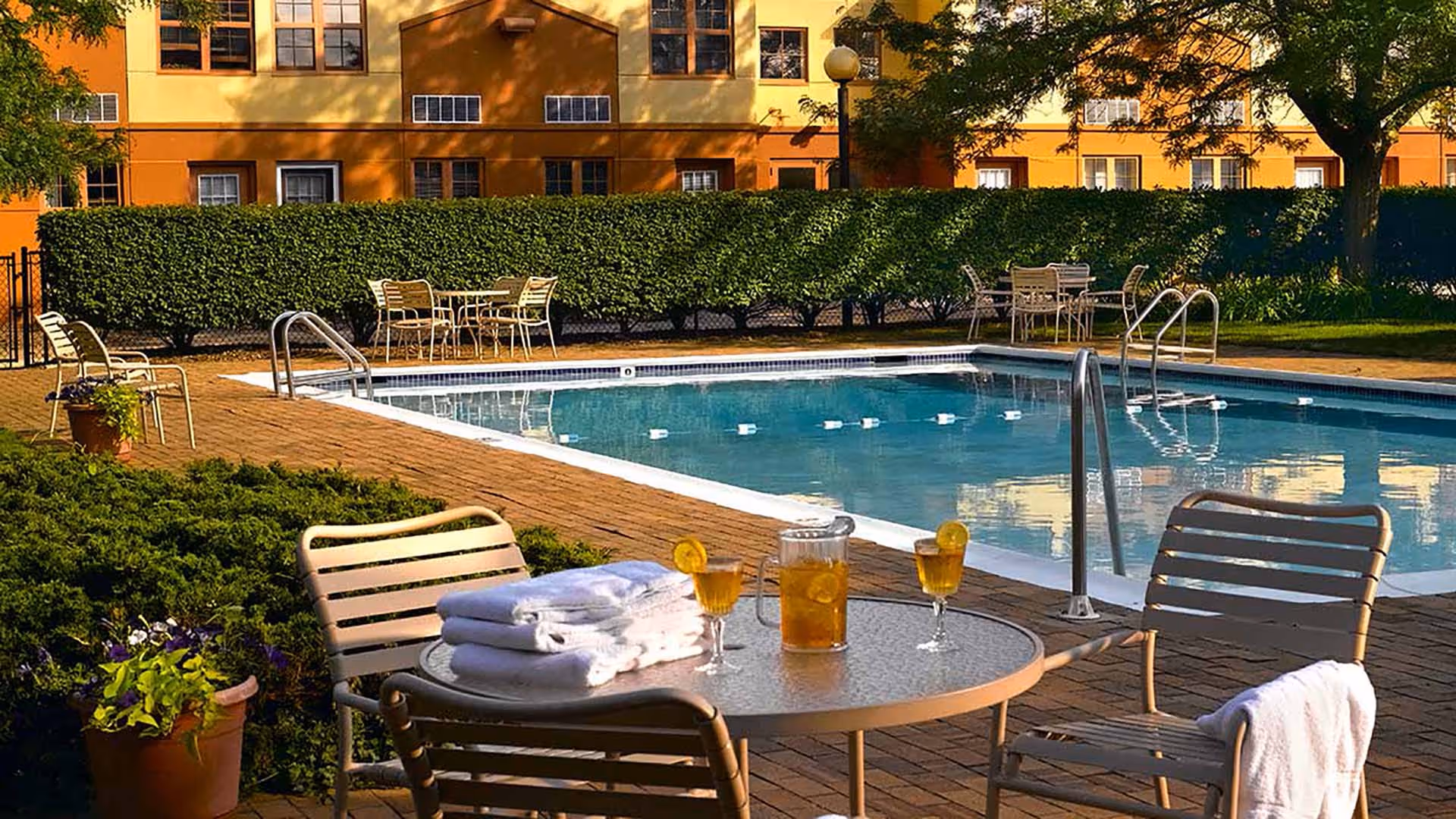 A sunny outdoor swimming pool with patio tables, lounge chairs, towels and drinks in front of a yellow residential building.