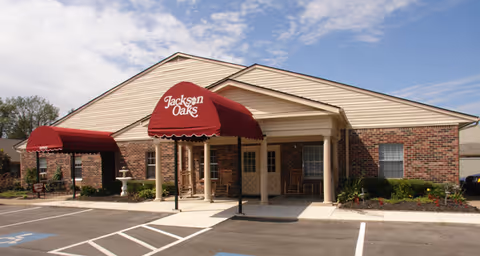 Exterior view of a single-story brick building with beige siding and two red awnings. The awning in the center reads 'Jackson Oaks'. There is a covered entrance with columns and rocking chairs on the porch. The parking lot in front has marked parking spaces, including handicapped spots.
