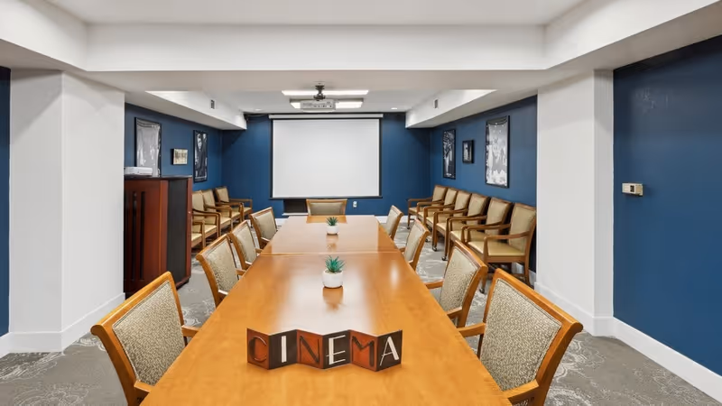A conference or meeting room with a long wooden table surrounded by chairs. The walls are painted blue with framed pictures hanging on them. At the far end of the room, there is a white projection screen and a ceiling-mounted projector. Small potted plants are placed on the table, and a decorative sign spelling 'CINEMA' is displayed in the center.