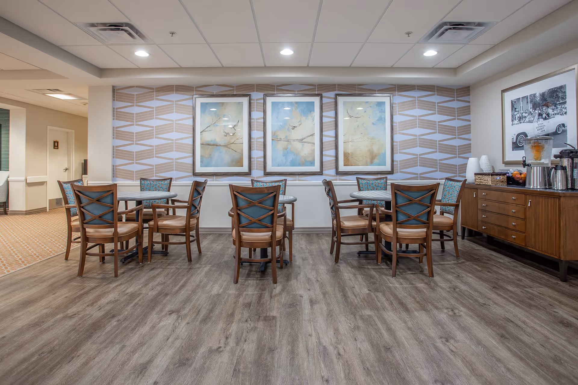 A dining area in a senior living facility with three round tables, each surrounded by four wooden chairs with cushioned seats and backs. The floor is wood-style laminate, and the walls feature geometric patterned wallpaper with three framed abstract paintings. To the right, there is a wooden sideboard with a beverage dispenser, coffee pots, and a framed black and white photograph above it. The ceiling has recessed lighting and air vents.