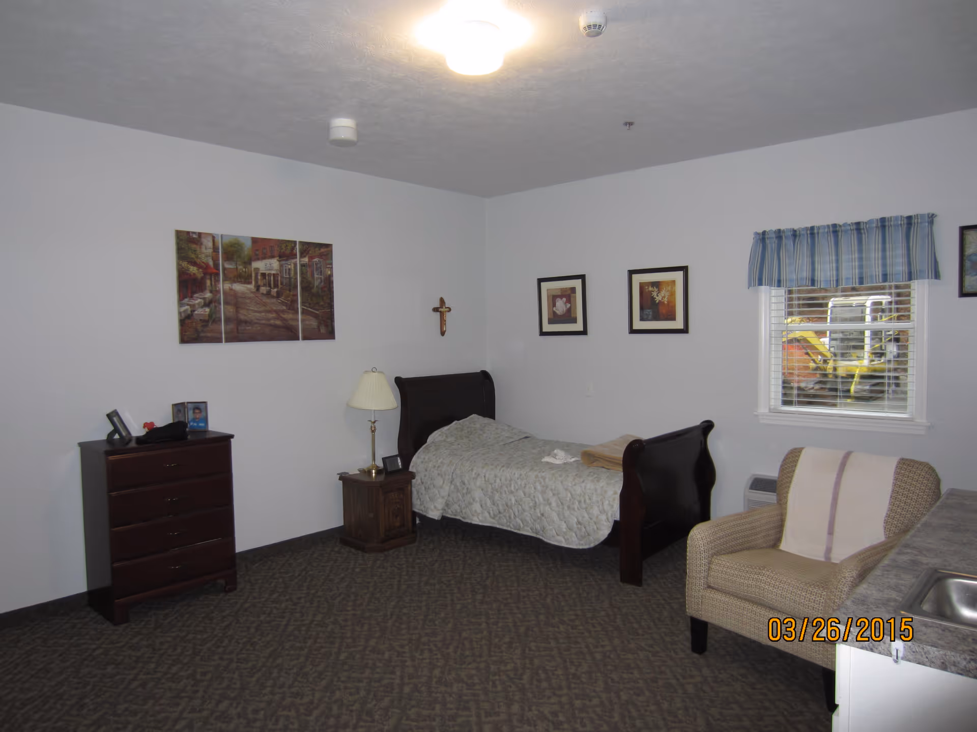 A simple bedroom with a single dark wooden bed covered with a light-colored quilt. Next to the bed is a wooden nightstand with a lamp and a small clock. On the left side of the room is a dark wooden dresser with framed photos on top. There are three framed pictures on the walls and a small window with blue plaid curtains showing construction equipment outside. A beige armchair with a striped blanket is near a countertop with a sink. The carpet is patterned and the walls are painted white.