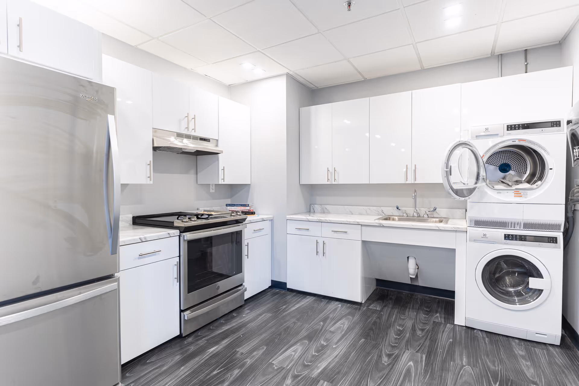Modern kitchen area with white cabinets, stainless steel refrigerator, stove, and a stacked washer and dryer unit with the dryer door open. The floor has a dark wood pattern, and the ceiling has recessed lighting.