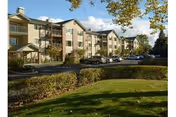 Multi-story residential building with balconies, landscaped lawn, and parked cars under a partly cloudy sky.