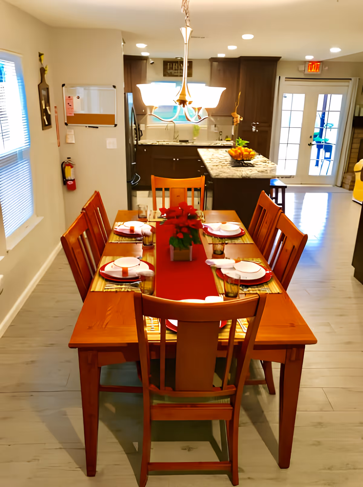 A dining area with a wooden table set for six people, featuring red placemats, white plates, and napkins. A red table runner with a small potted plant is centered on the table. In the background, there is a kitchen with dark wood cabinets, a granite countertop island with a bowl of fruit, and a chandelier hanging above the dining table. A window with blinds and a fire extinguisher are visible on the left wall, and glass doors leading outside are seen in the background.