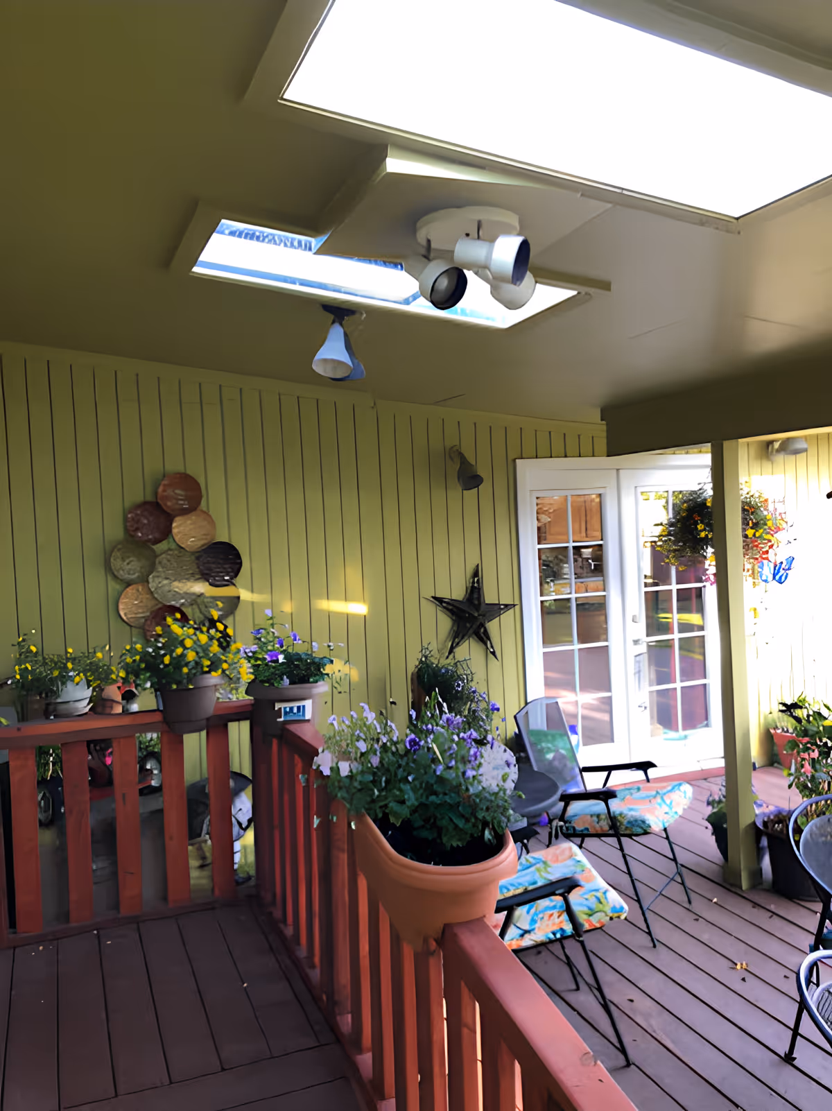Covered outdoor patio area with wooden flooring and red railing, decorated with potted flowers and plants. There are two chairs with colorful cushions and a small round table. The walls are painted yellow-green with decorative items including a metal star and circular wall art. Skylights and ceiling lights are visible above, and glass double doors lead inside.