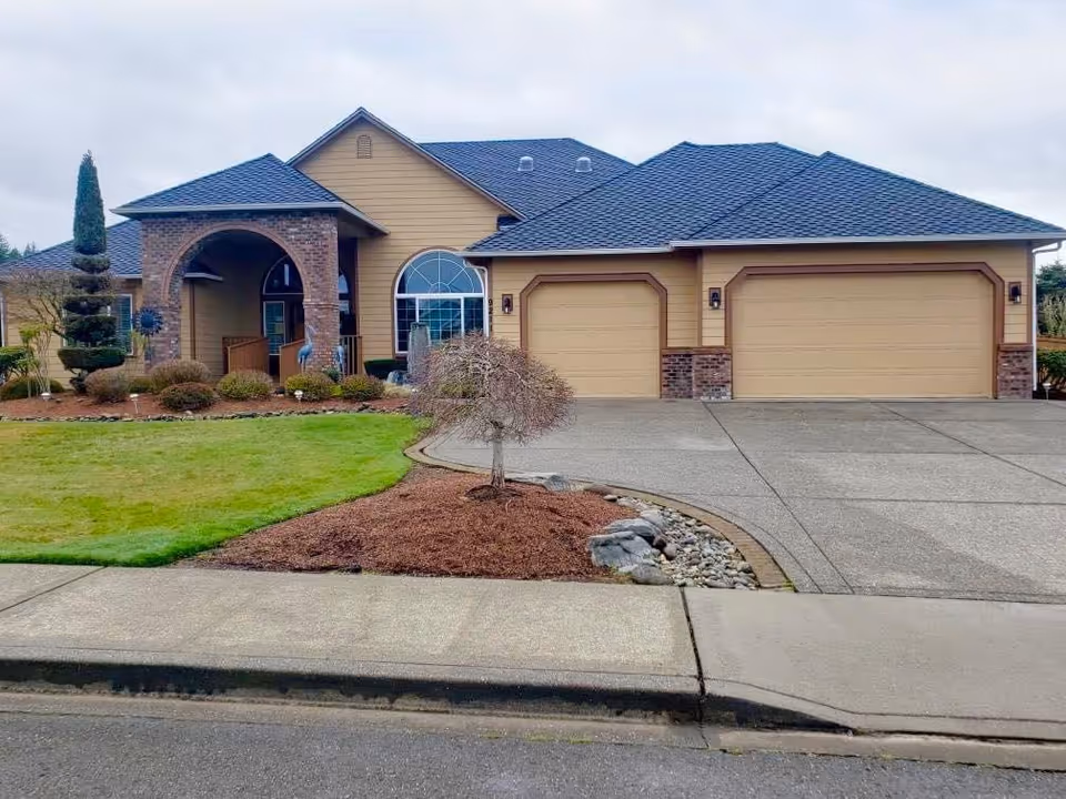 Front exterior view of a single-story residential building with a three-car garage, a covered entrance with brick pillars, manicured lawn, and landscaped garden beds.