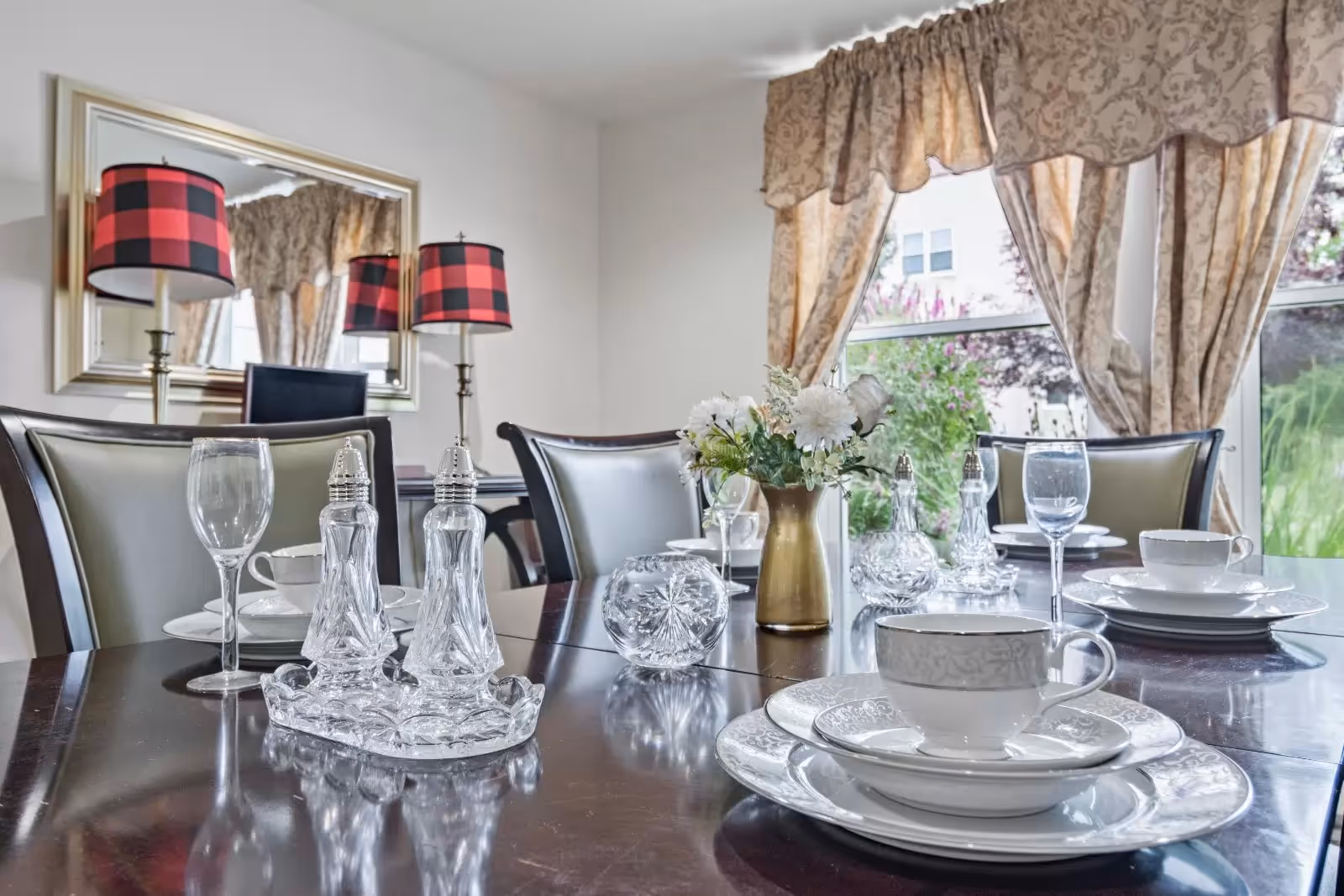 A dining table set with white china plates, cups, and wine glasses. There is a gold vase with white flowers in the center, along with crystal salt and pepper shakers. The room has beige patterned curtains and a large mirror on the wall reflecting two lamps with red and black checkered lampshades.