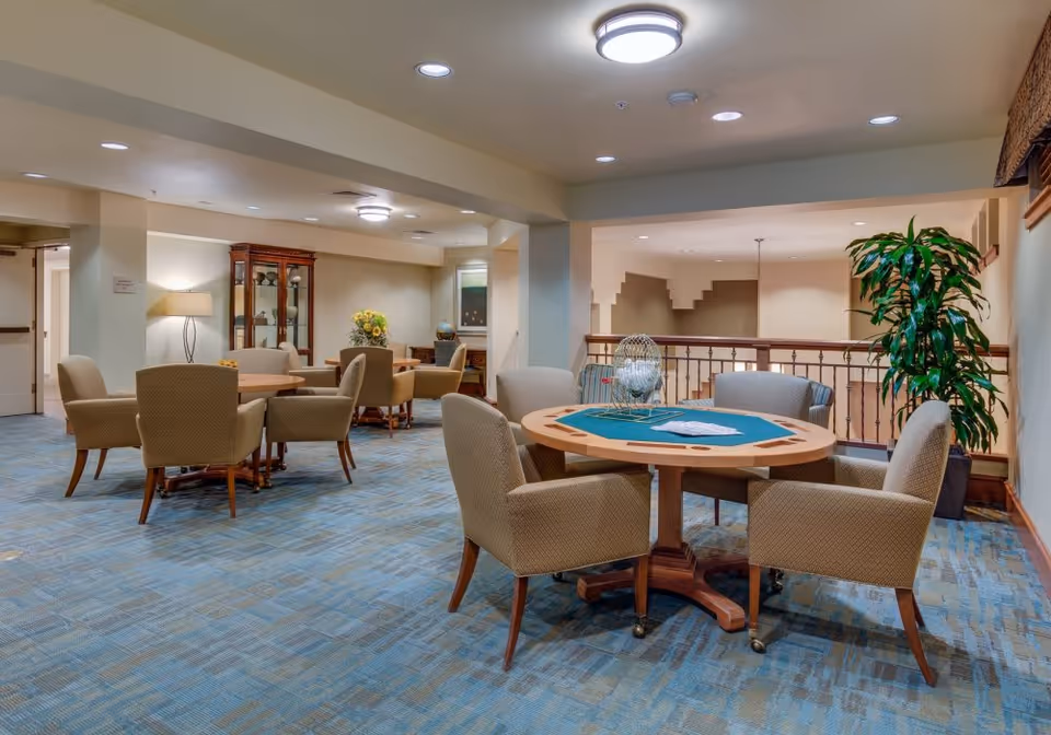 A well-lit common area in a senior living facility featuring multiple round tables with comfortable upholstered chairs arranged around them. One table has a bingo cage and cards, suggesting a recreational space. The room has blue patterned carpet, beige walls, and ceiling lights. There is a large potted plant near a railing overlooking a lower level, and a wooden cabinet with decorative items in the background.