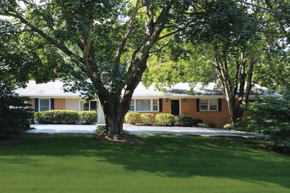 A single-story brick building partially obscured by large leafy trees and bushes, with a well-maintained lawn in the foreground and a paved driveway leading to the entrance.