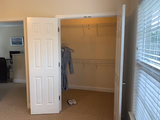 An open closet with white double doors showing empty wire shelves and a few hangers holding a gray robe. A pair of slippers is on the carpeted floor inside the closet. To the right, there is a window with white blinds partially open, and to the left, part of a living room with a sofa and a framed picture on the wall is visible.