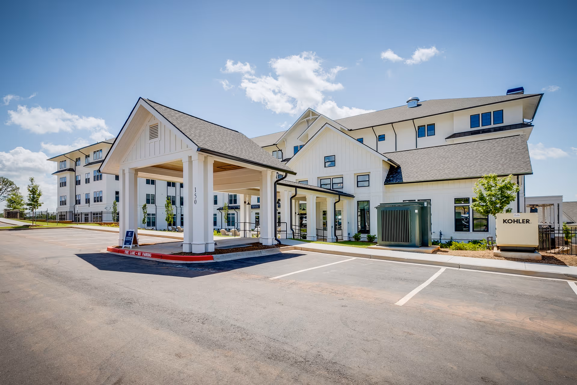 Exterior view of a modern senior living facility building with white siding and a covered entrance. The building has multiple floors with numerous windows and a clear blue sky in the background. There is a parking area in front and some landscaping with small trees and shrubs.