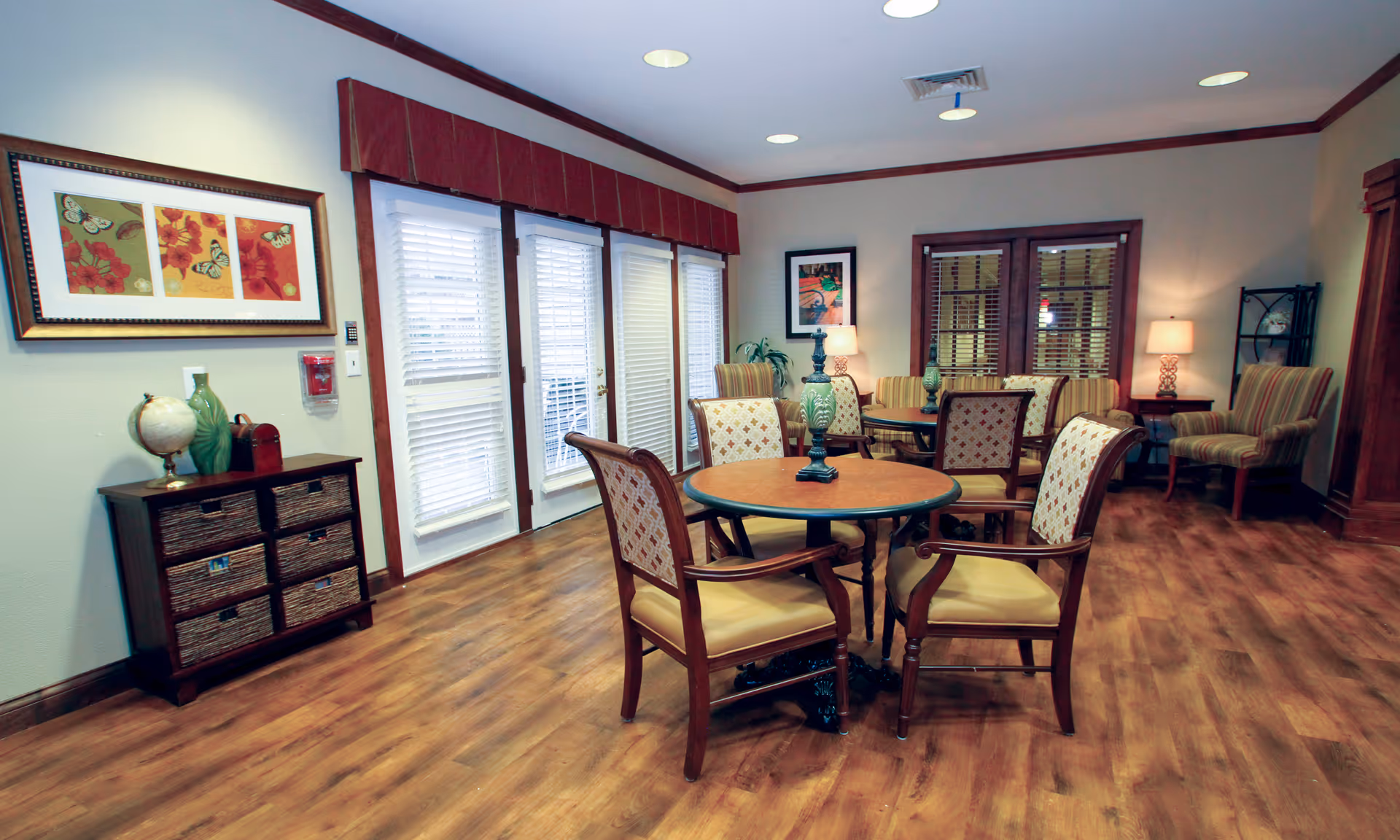 Spacious senior living lounge with round table, chairs, and warm wood flooring.