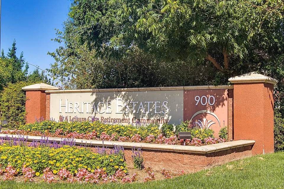 Outdoor entrance sign for Heritage Estates, a retirement community, surrounded by colorful flowers and greenery under a clear blue sky.